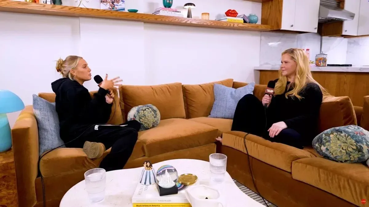 Two women in black hold handheld mics on an L-shaped tan sectional in an open apartment living room beside a kitchen.